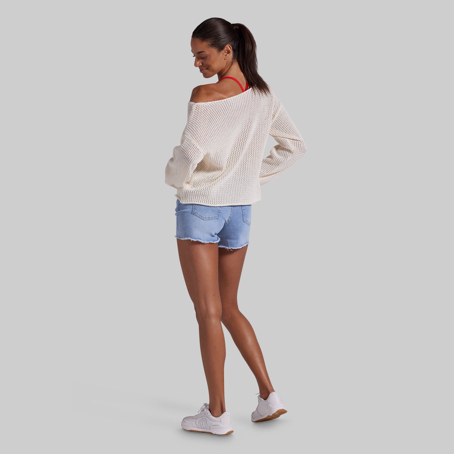 A woman modeling an off-the-shoulder cream knit sweater, light blue denim shorts, and white sneakers, shown from a side-back angle against a solid gray background.