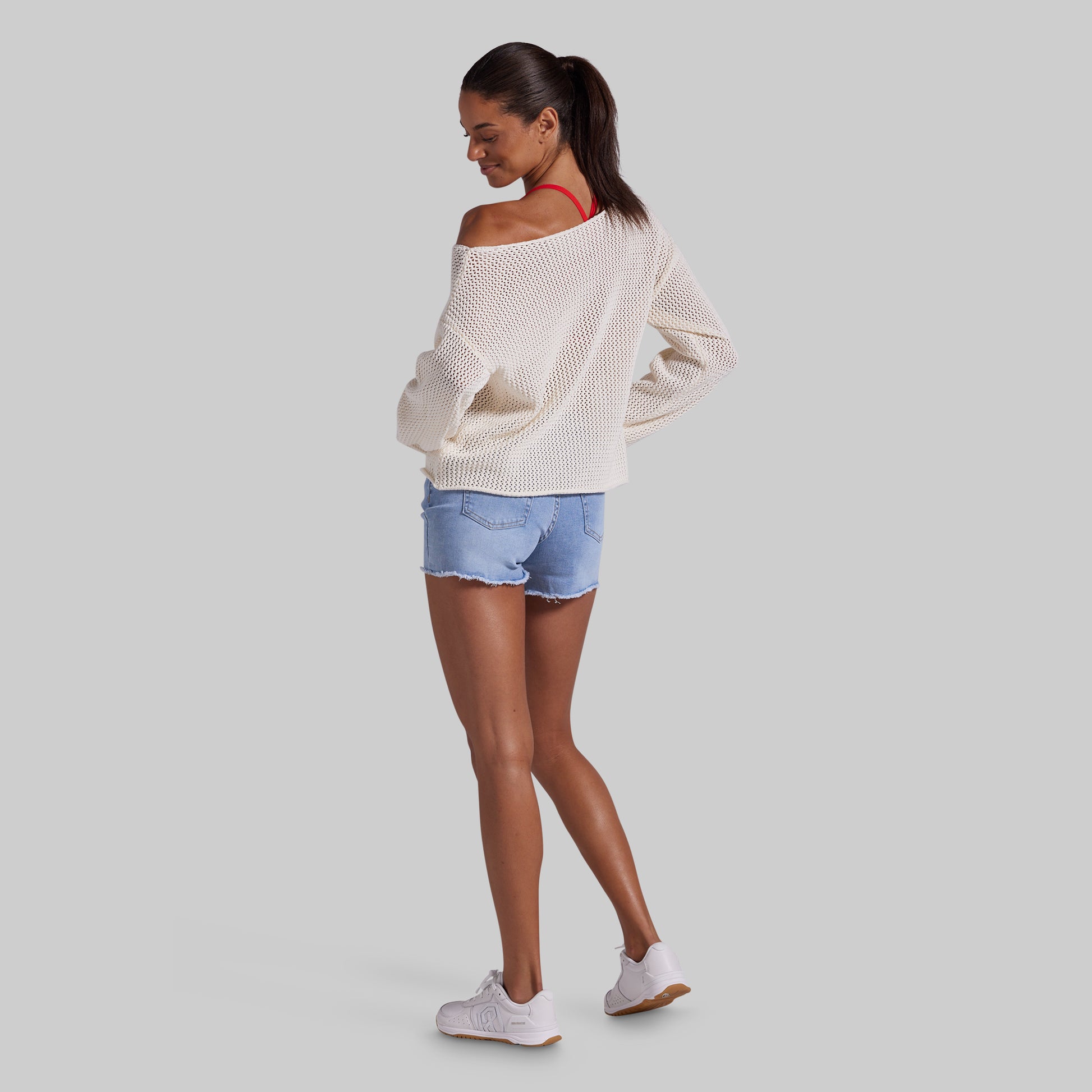 A woman modeling an off-the-shoulder cream knit sweater, light blue denim shorts, and white sneakers, shown from a side-back angle against a solid gray background.