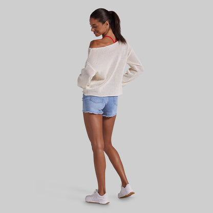 A woman modeling an off-the-shoulder cream knit sweater, light blue denim shorts, and white sneakers, shown from a side-back angle against a solid gray background.