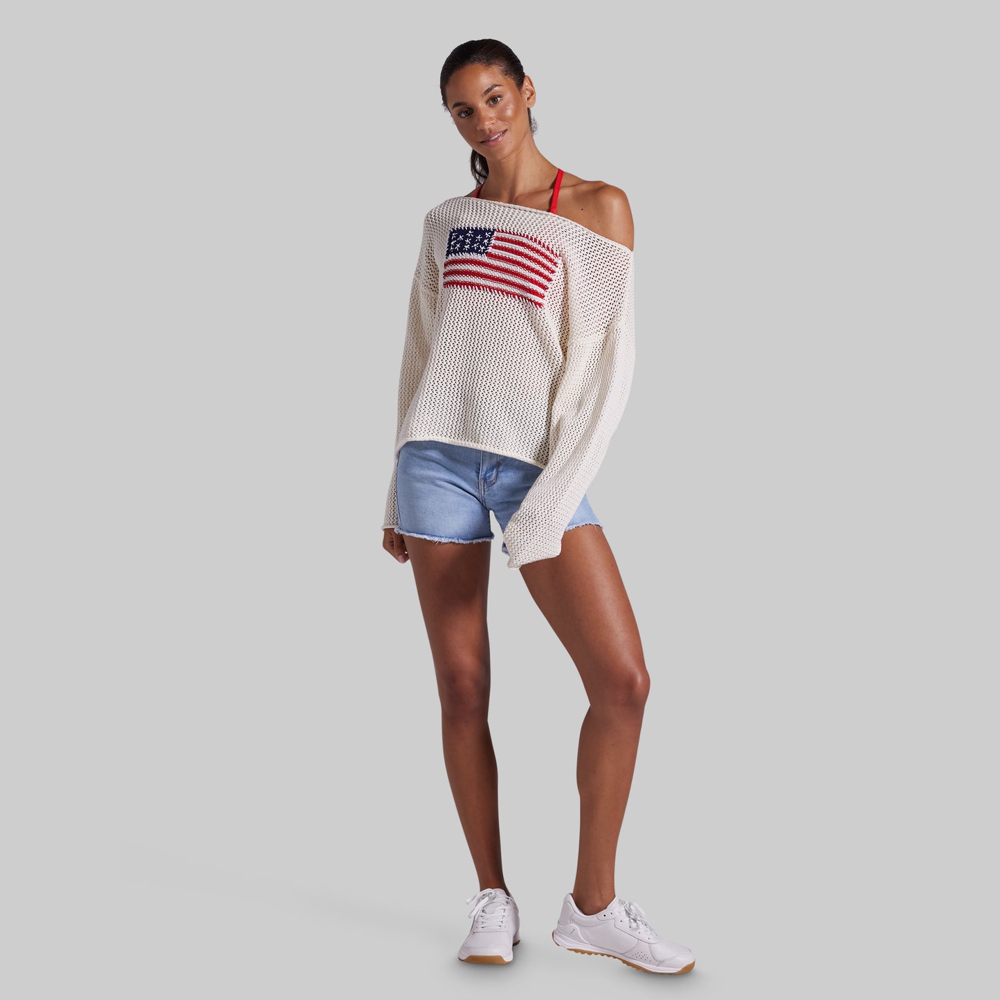 A woman modeling a white off-the-shoulder knit sweater with a red, white, and blue American flag graphic on the chest, paired with denim shorts and white sneakers, against a solid gray background.