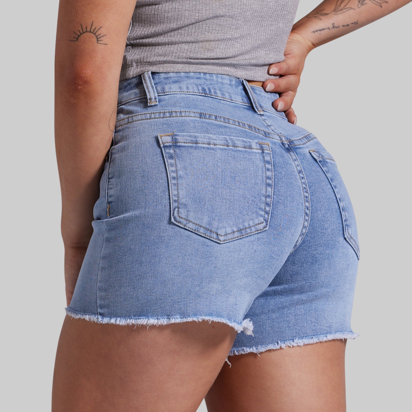 Rear view of a person wearing light blue denim cutoff shorts with a frayed hem, paired with a gray ribbed top, against a light gray studio background.