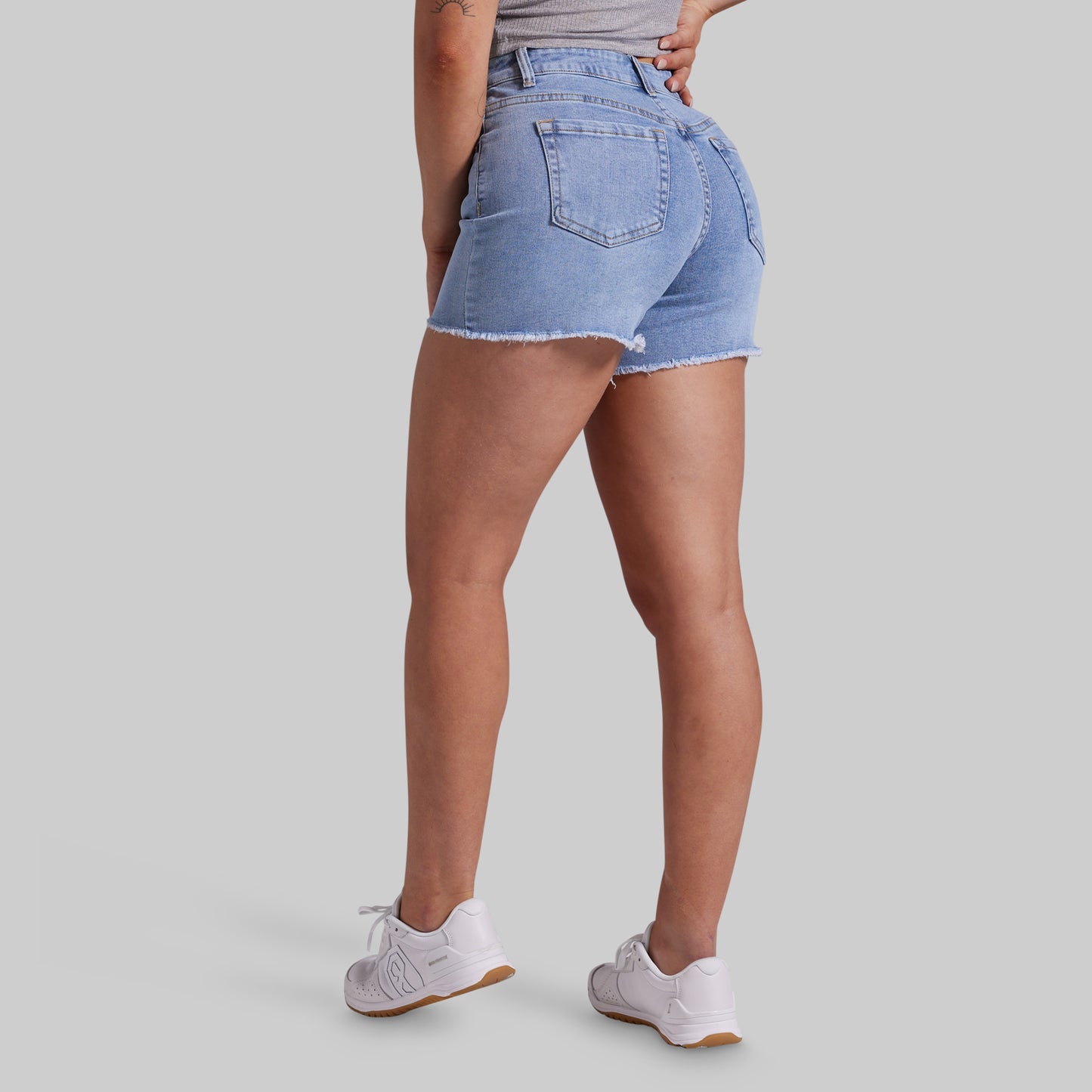 A model from the back wearing light-wash blue denim shorts with a frayed hem, paired with white sneakers, against a solid gray studio background.