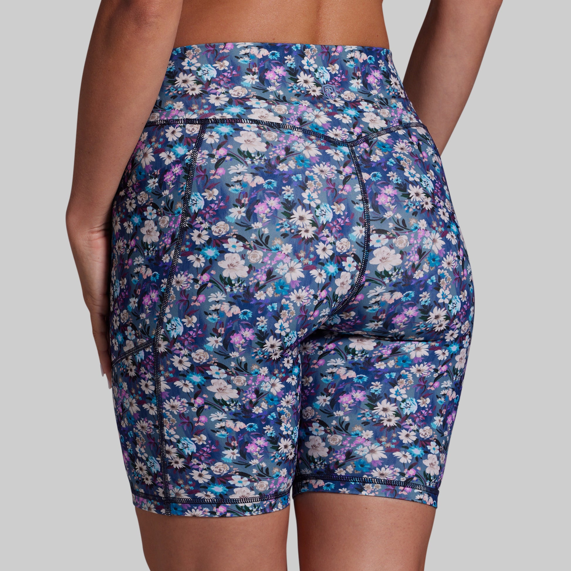 Rear view of a person wearing high-waisted floral biker shorts with a blue, purple, and white daisy pattern against a solid gray background.