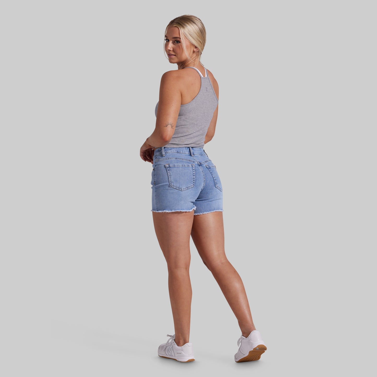 A woman modeling light-wash denim shorts, a gray tank top, and white sneakers, shown from a rear three-quarter angle against a solid gray studio background.
