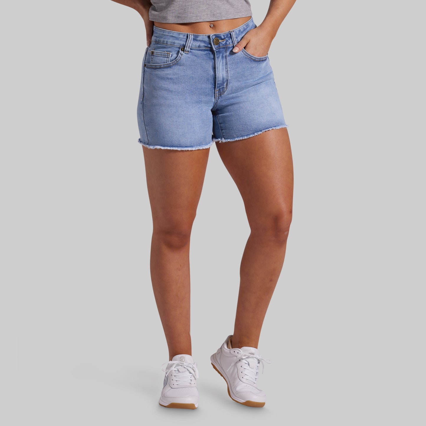 A model wearing light-wash blue denim shorts with a frayed hem, paired with white sneakers and a gray crop top, shown from the waist down on a solid gray background.