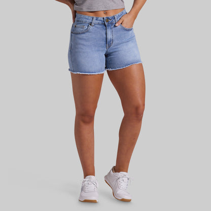 A model wearing light-wash blue denim shorts with a frayed hem, paired with white sneakers and a gray crop top, shown from the waist down on a solid gray background.