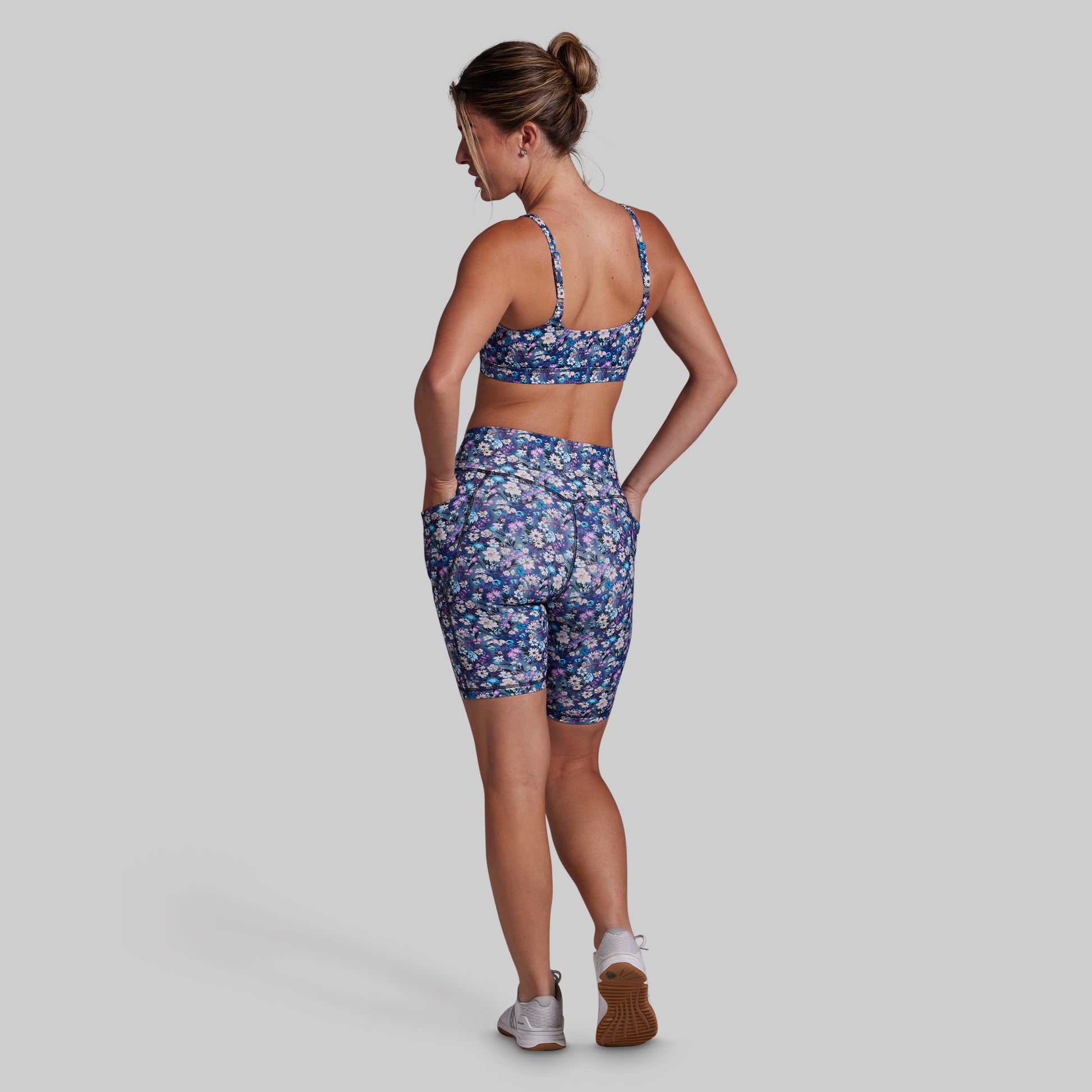 A woman is shown from the back wearing a matching athletic set consisting of a blue floral sports bra and biker shorts, set against a solid gray background.
