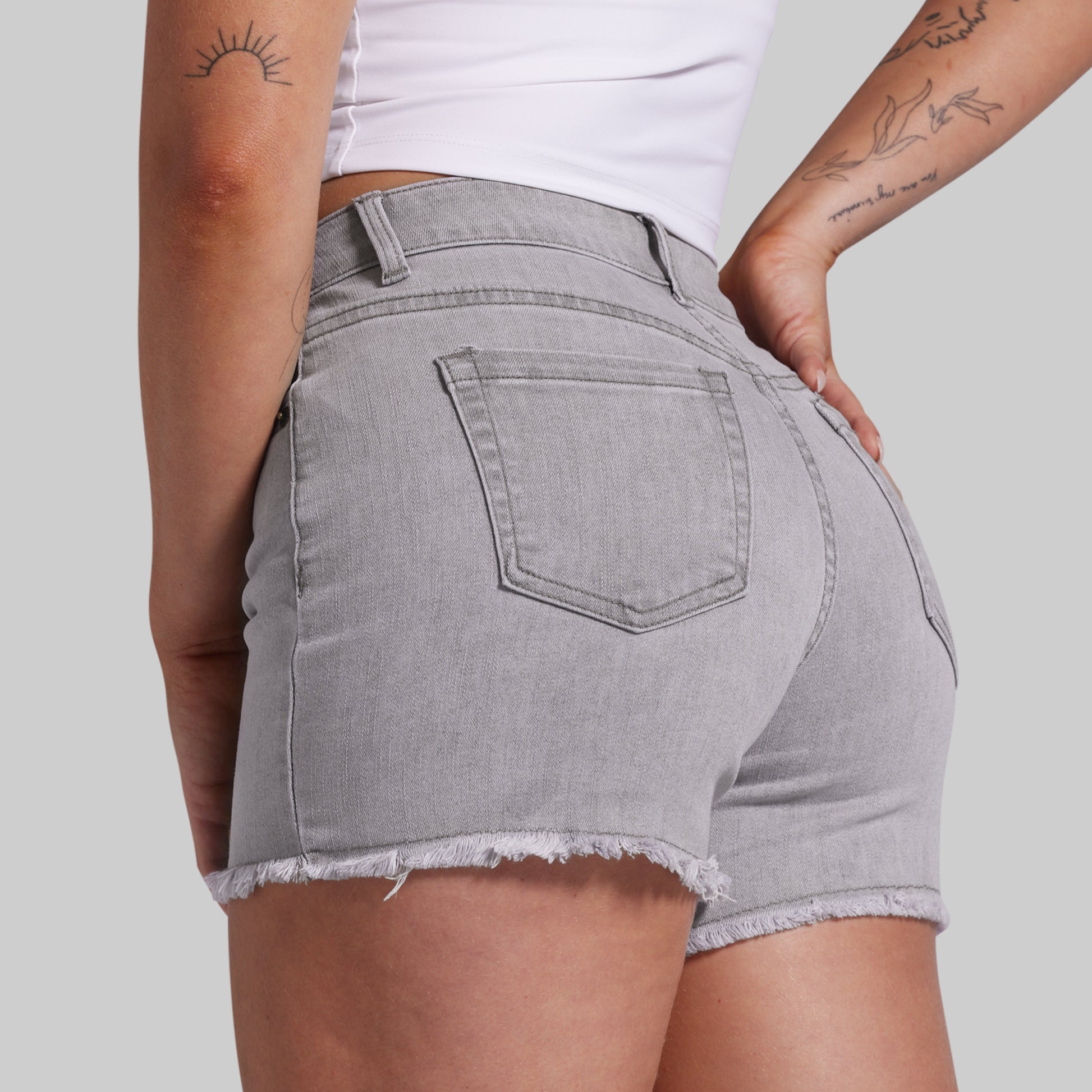 A close-up back view of a model wearing light gray denim shorts with a frayed hem and a back pocket, paired with a white top against a solid gray background.
