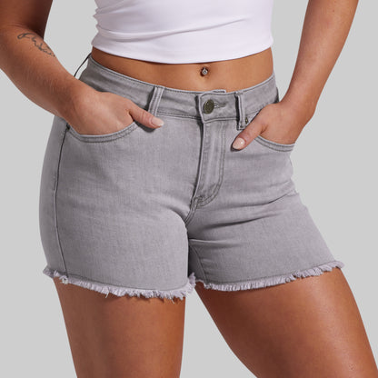 A close-up of a model wearing light gray denim shorts with a raw, frayed hem and classic five-pocket styling, paired with a white crop top against a solid gray background.