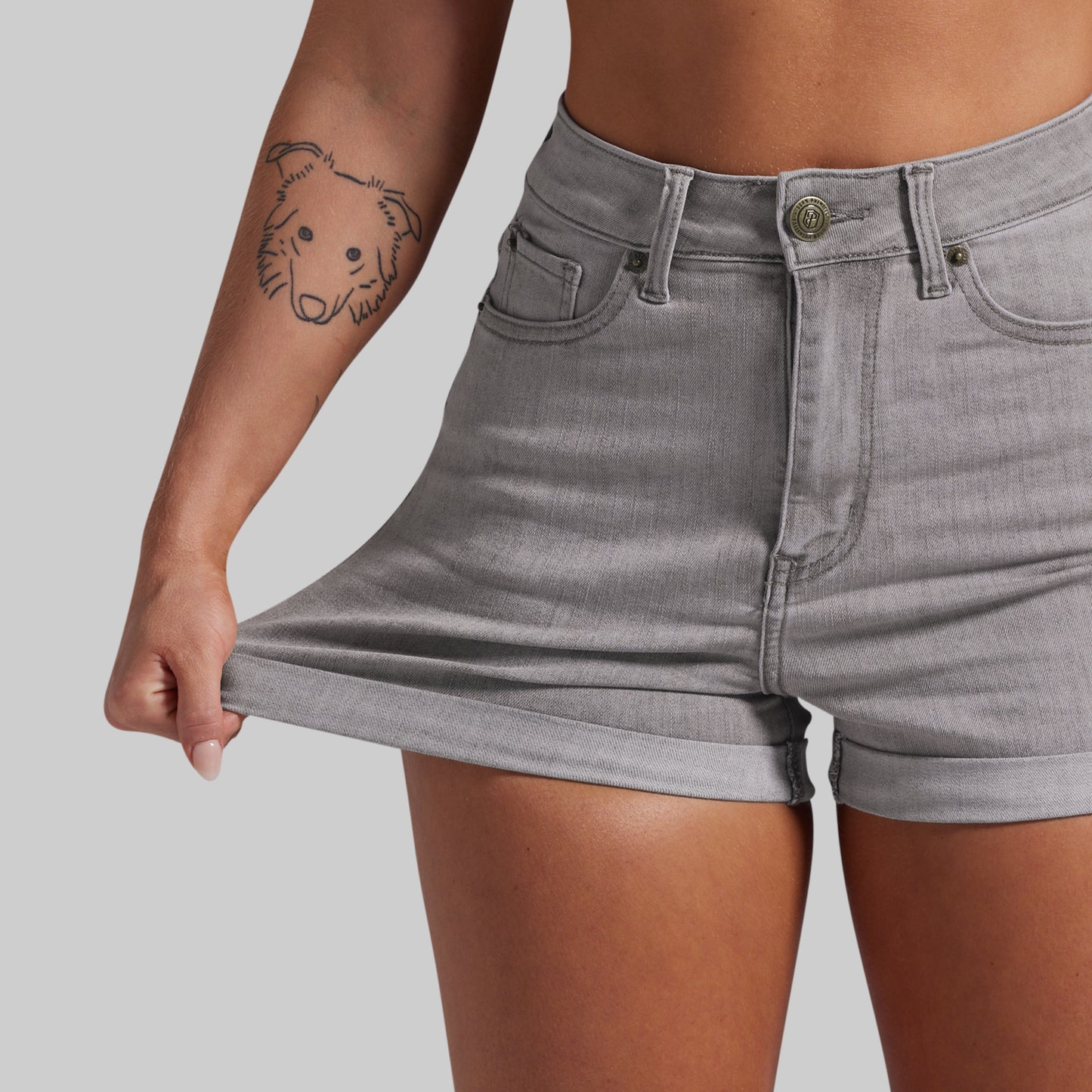 A close-up of a model wearing light gray denim shorts, pulling the hem slightly to show the fabric stretch. 