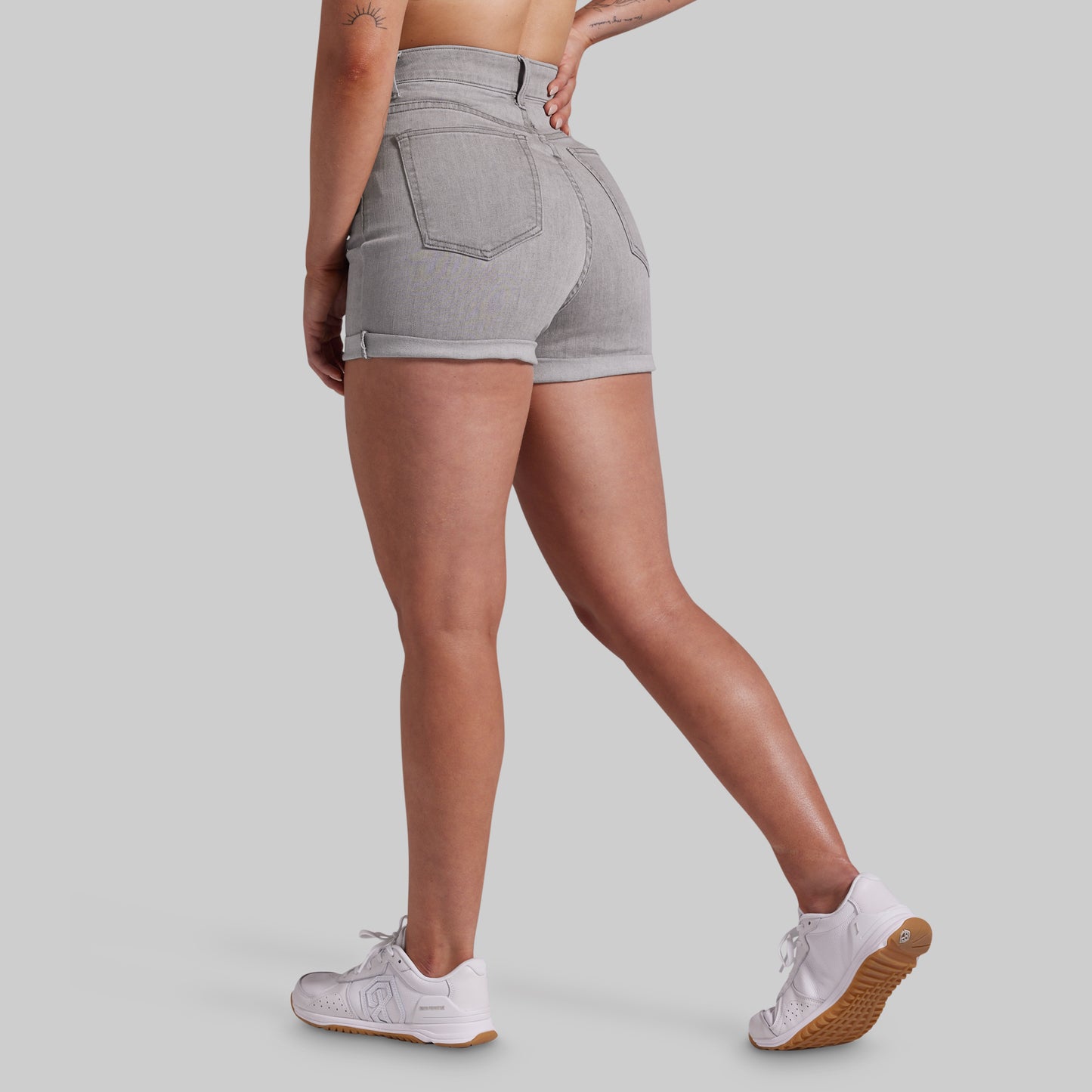 A model shown from the back wearing light gray high-waisted denim shorts and white sneakers, set against a solid light gray studio background.