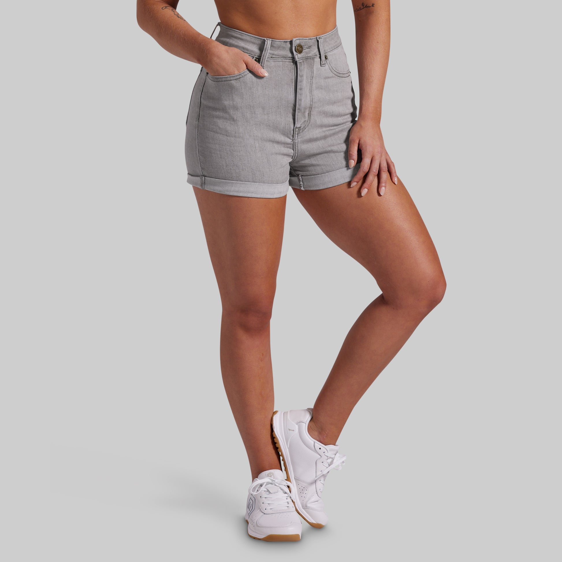 A model wearing light gray denim shorts with rolled hems and white sneakers, shown from the waist down against a solid gray background.