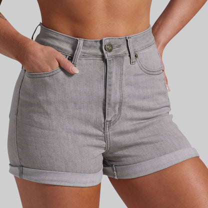 A close-up of a model wearing light gray denim shorts with a rolled hem, styled with hands in the pockets, against a solid gray background.