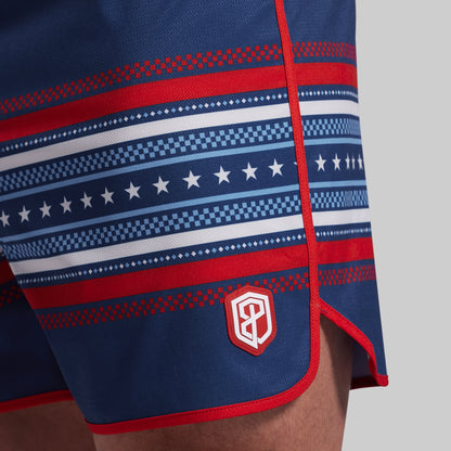 Close-up of navy blue boatf shorts with red and white horizontal patterned stripes, a small shield logo patch near the hem, and red trim, shown on a model against a light gray background.