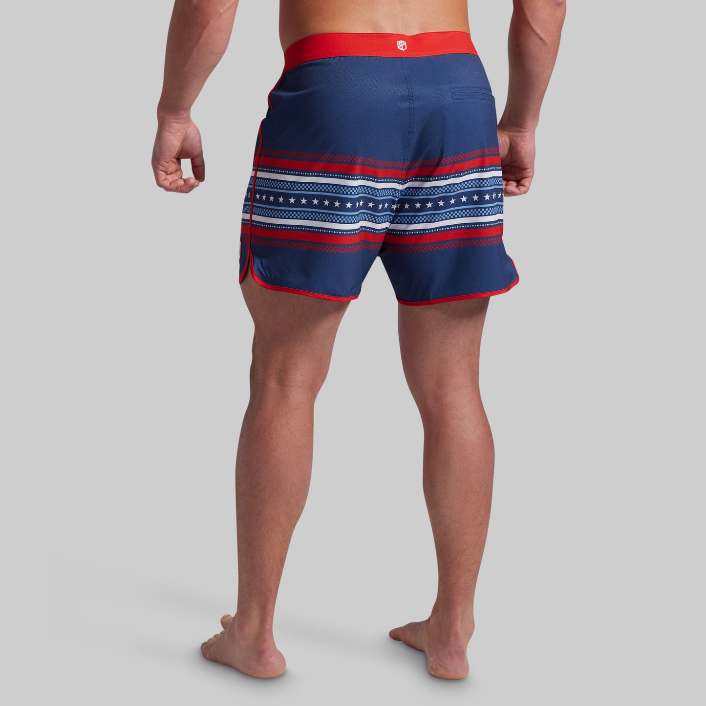 Rear view of a man wearing navy blue swim trunks with red and white horizontal stripes and a red waistband, against a solid gray background.