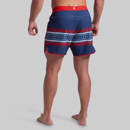 Rear view of a man wearing navy blue swim trunks with red and white horizontal stripes and a red waistband, against a solid gray background.