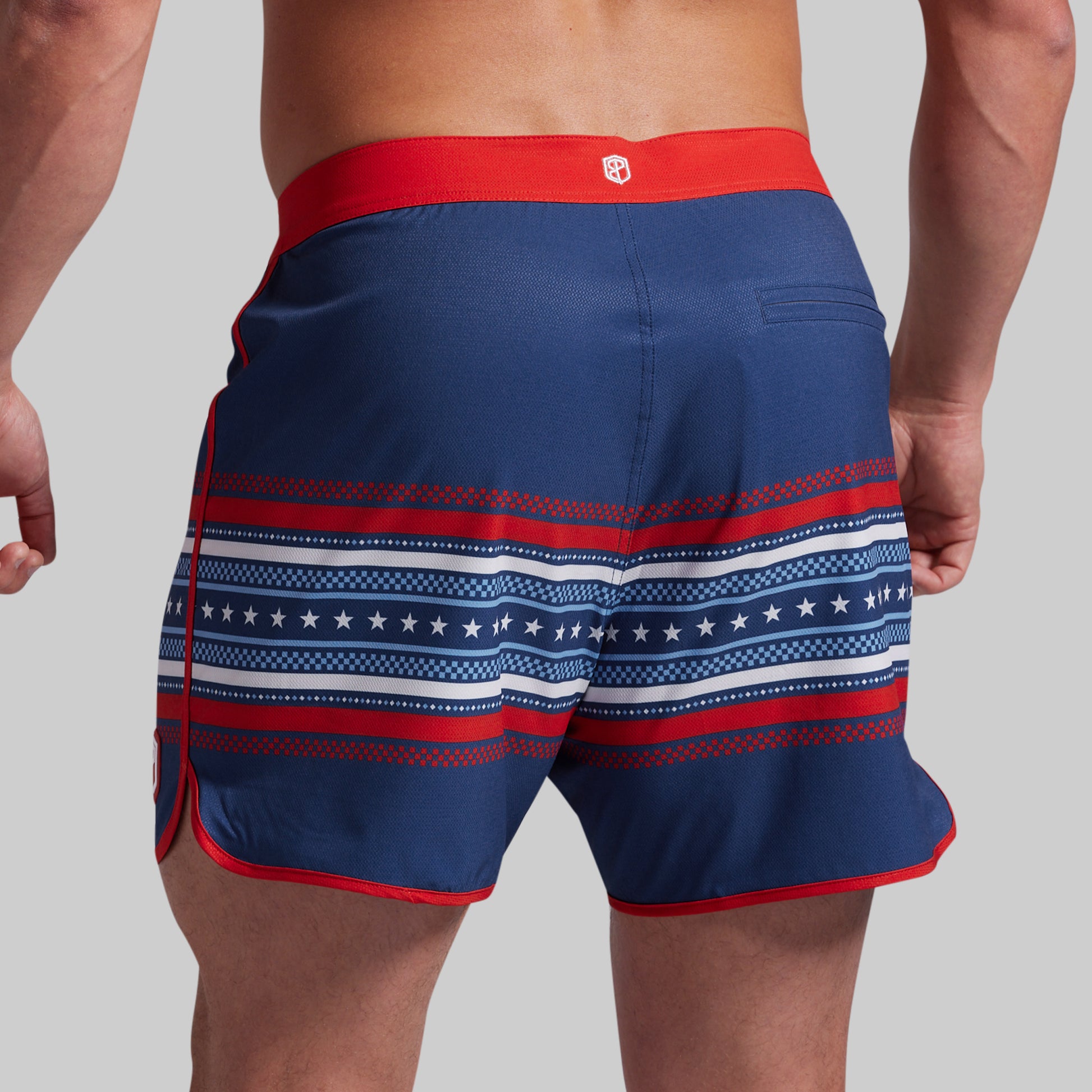 A close-up back view of a model wearing navy blue board shorts with red and white patriotic patterns, including stars and stripes, on a solid gray background.