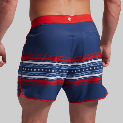 A close-up back view of a model wearing navy blue board shorts with red and white patriotic patterns, including stars and stripes, on a solid gray background.