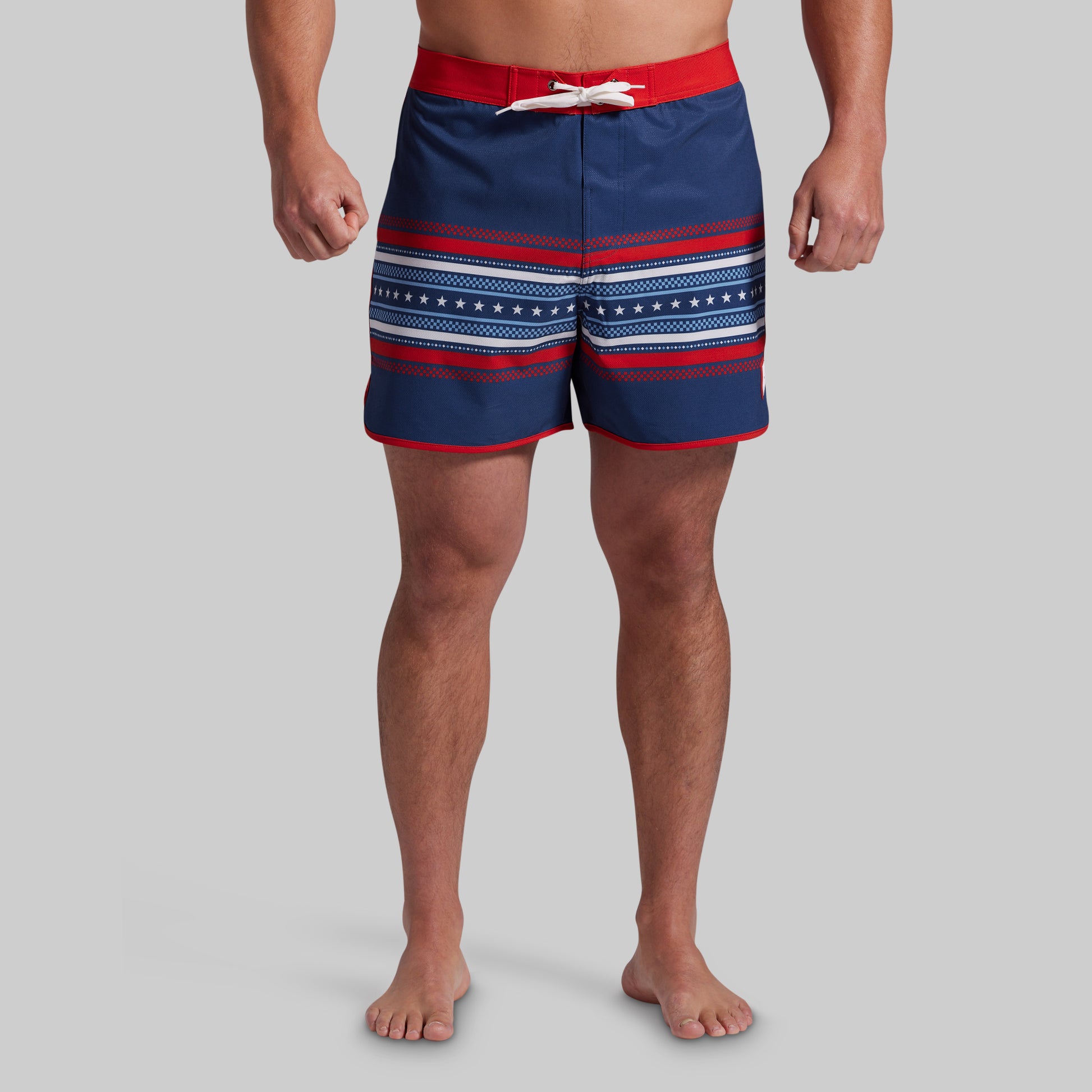 A man wearing navy blue board shorts with red and white horizontal stripes and a red waistband, standing against a solid gray background.