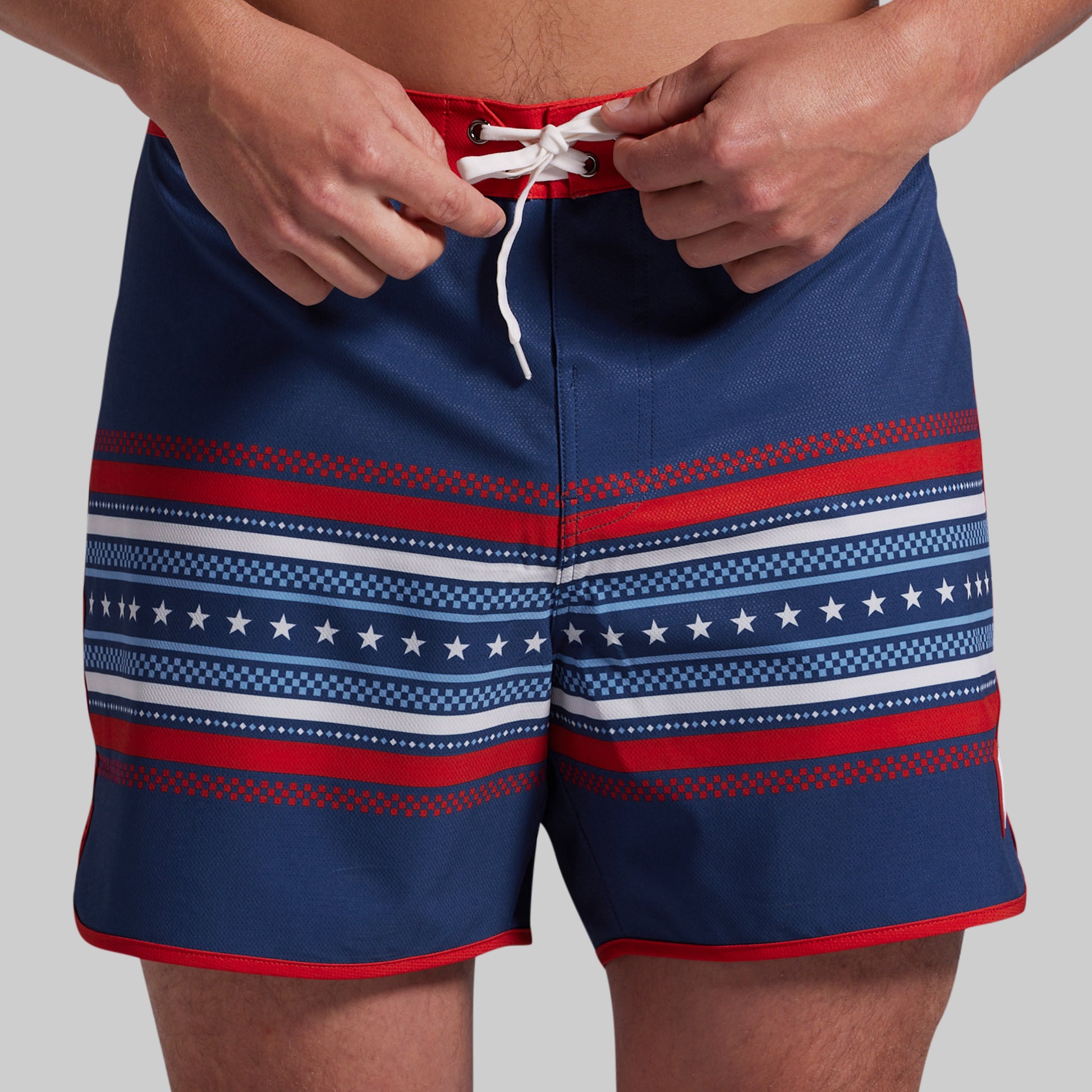 A close-up of a person wearing navy board shorts with red and white horizontal patterned stripes and a white drawstring, shown from the waist to mid-thigh against a light gray studio background.