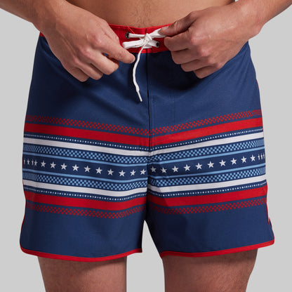 A close-up of a person wearing navy board shorts with red and white horizontal patterned stripes and a white drawstring, shown from the waist to mid-thigh against a light gray studio background.