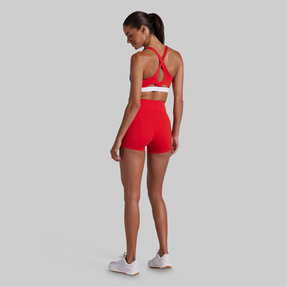 A woman wearing a matching red activewear set consisting of a sports bra and high-waisted shorts, shown from a rear three-quarter view against a solid gray background.
