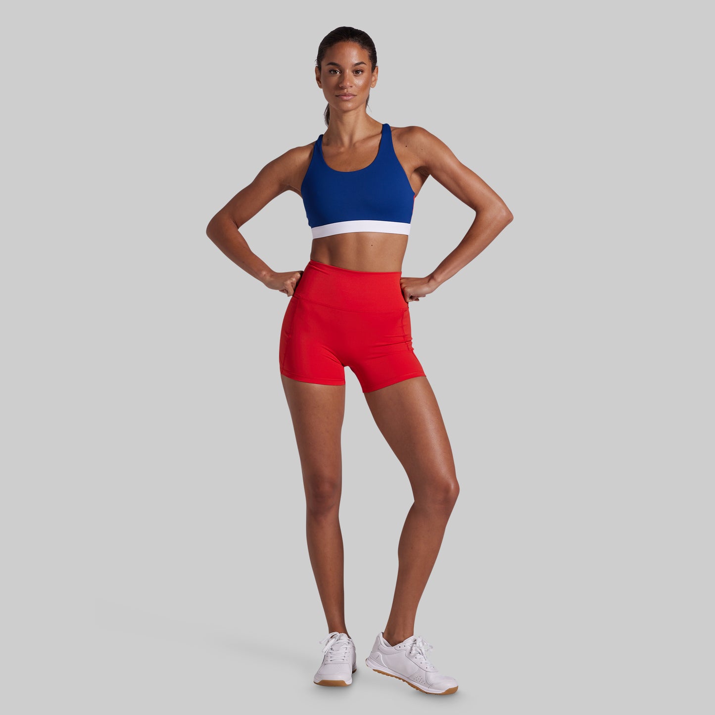 A woman posing in a blue and white sports bra and bright red athletic shorts against a solid light gray background.