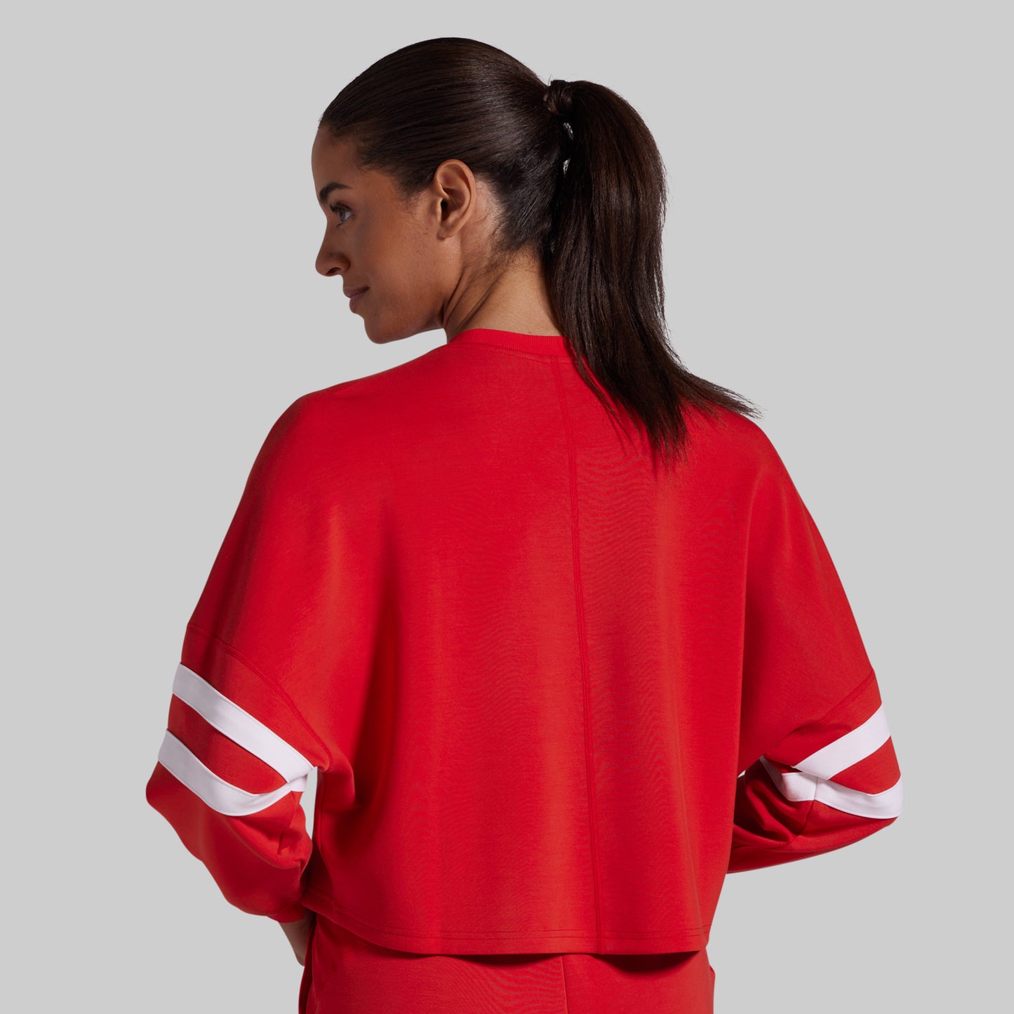 A woman shown from the back wearing a bright red cropped athletic sweatshirt with white double stripes on the sleeves, against a solid gray background.