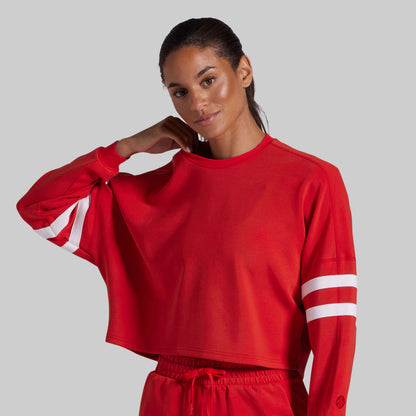 A woman modeling a bright red cropped long-sleeve sweatshirt with white double stripes on both sleeves, paired with matching red shorts, against a solid gray background.