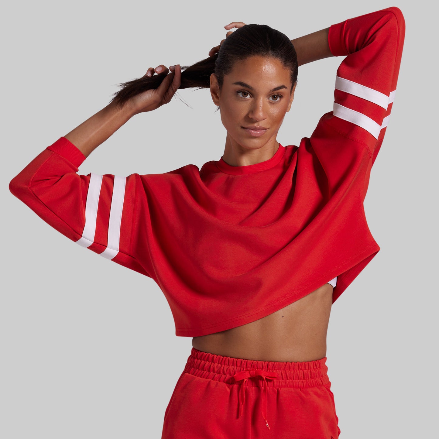A woman modeling a bright red cropped long-sleeve sweatshirt with white stripe details on the sleeves, paired with matching red shorts, against a solid gray background.