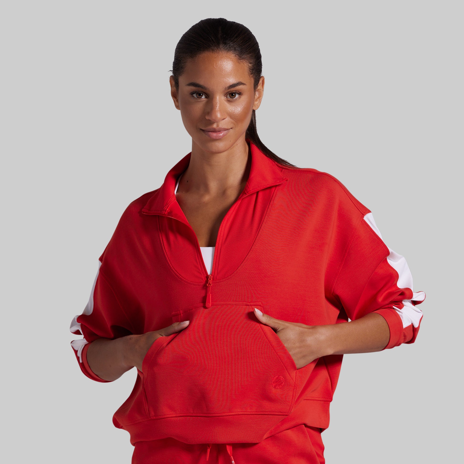 A woman modeling a bright red athletic quarter zip-up jacket with white sleeve stripes, posed against a solid light gray background.