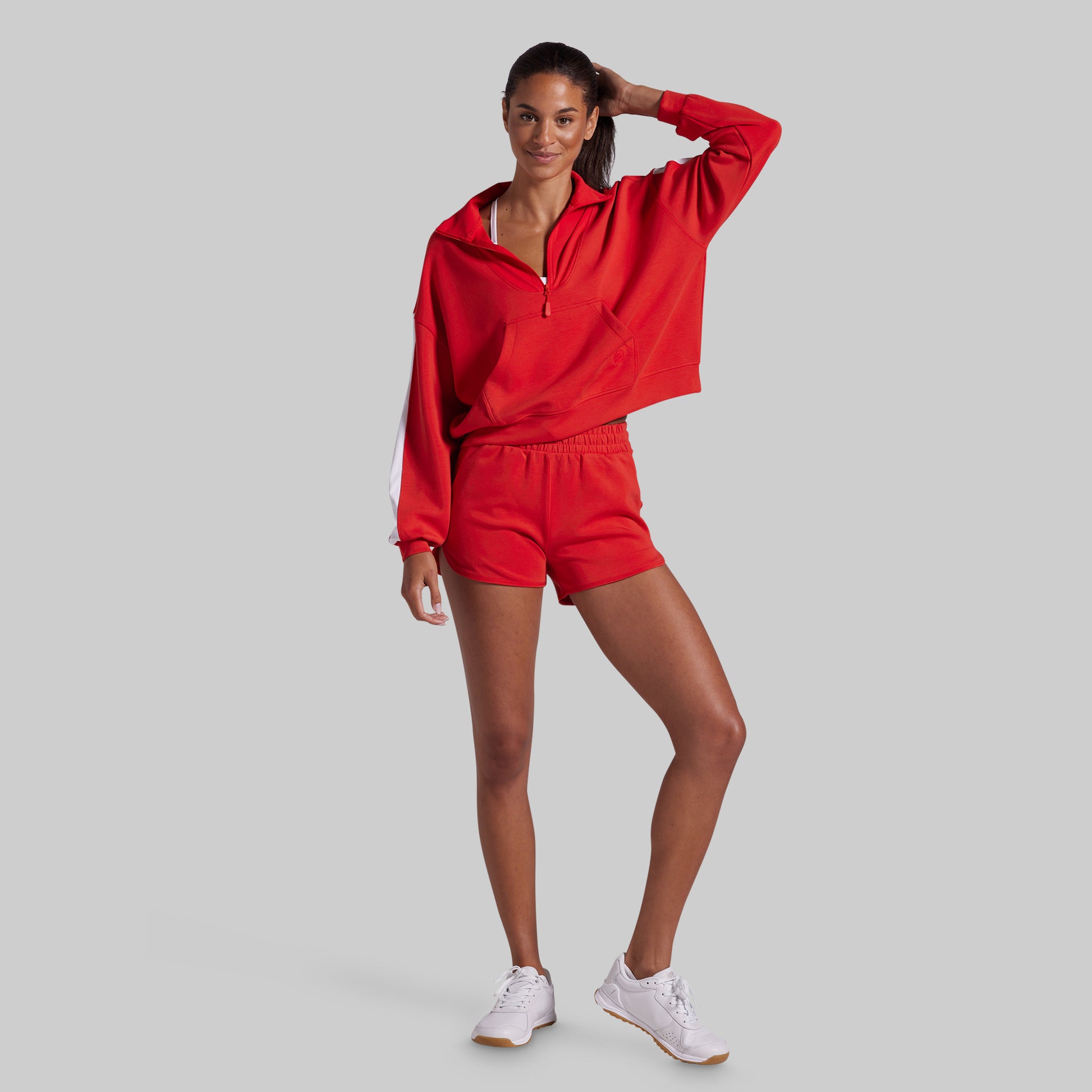 A woman modeling a matching red athletic set consisting of a cropped zip-up hoodie and shorts, paired with white sneakers, against a solid gray background.