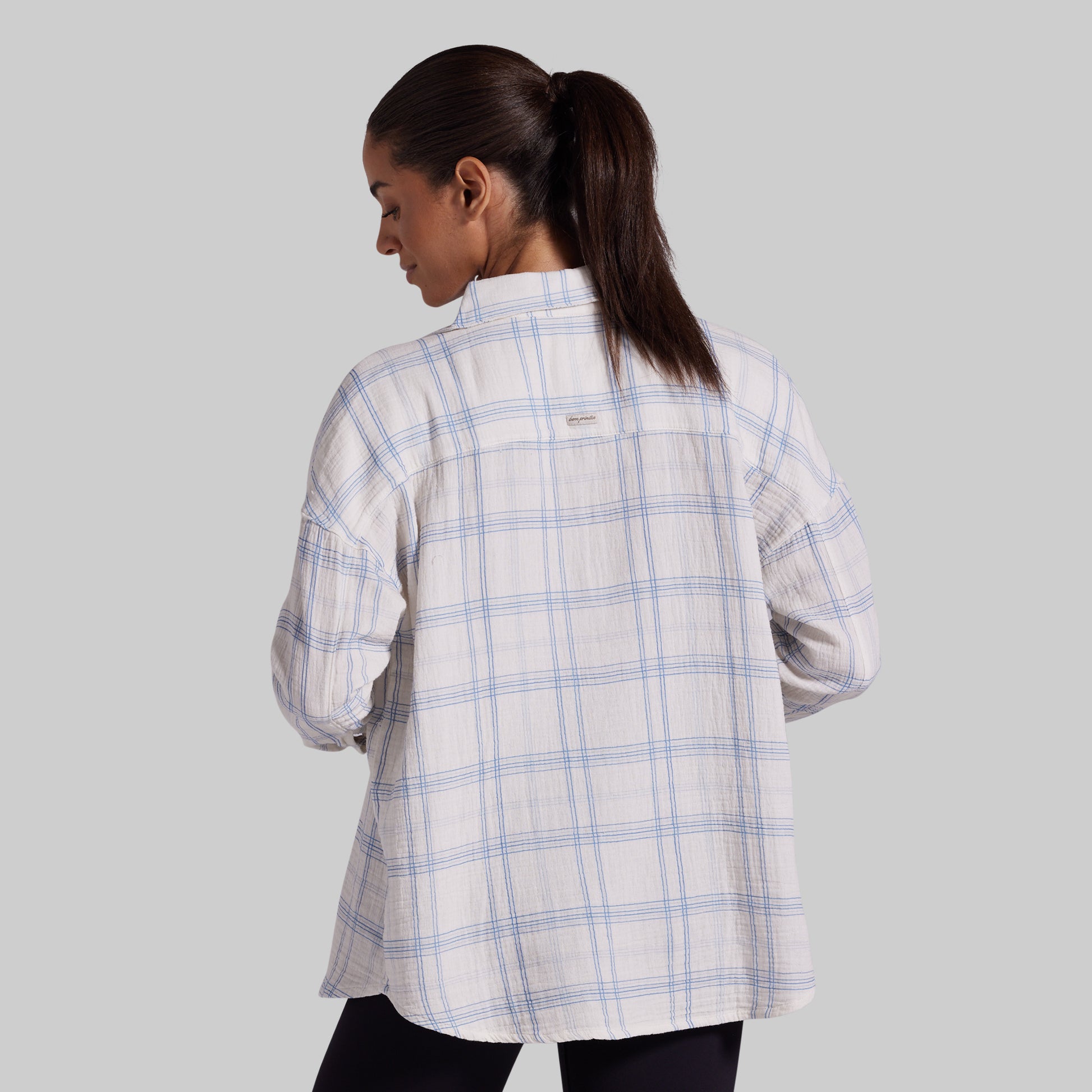 A woman shown from the back wearing a white long-sleeve plaid button-up shirt with thin blue lines, paired with black pants, against a solid gray studio background.
