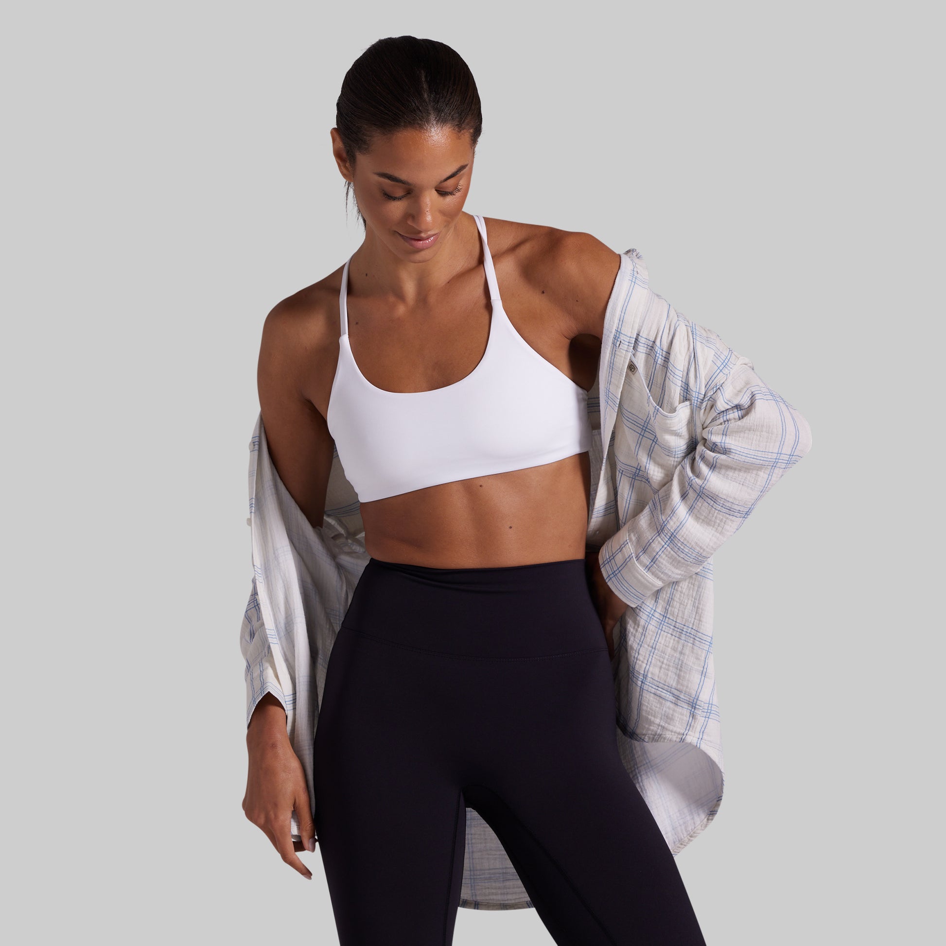 A woman modeling a white sports bra and black leggings, with an open light blue and white plaid flannel shirt draped off her shoulders, against a solid gray background.