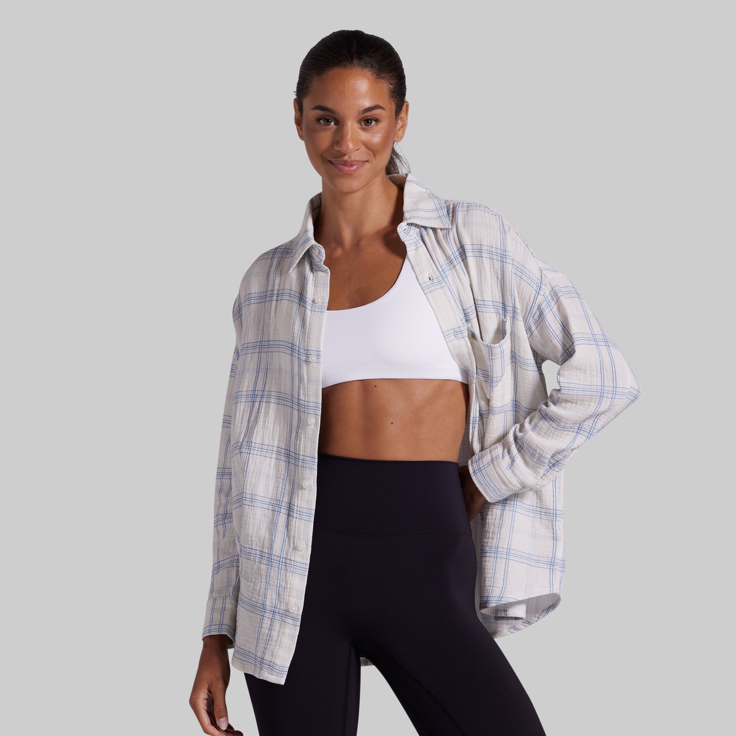 A woman modeling a white and light blue plaid button-up shirt over a white sports bra and black leggings, set against a solid gray studio background.