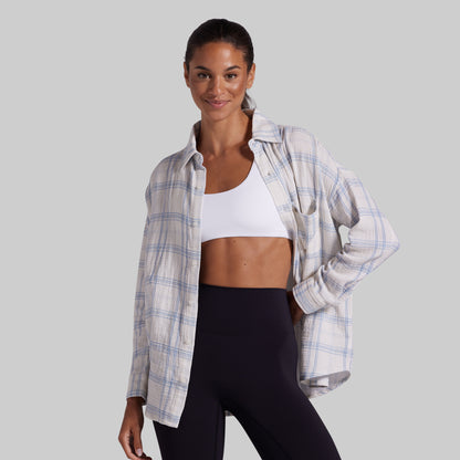 A woman modeling a white and light blue plaid button-up shirt over a white sports bra and black leggings, set against a solid gray studio background.