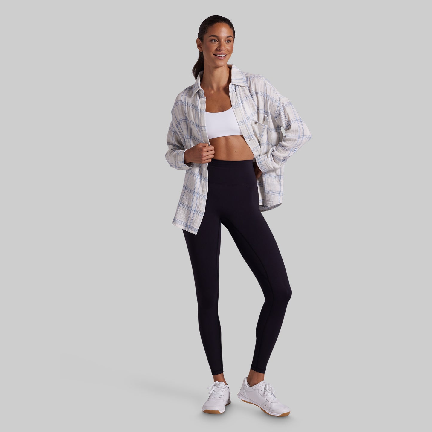 A woman modeling a pair of black high-waisted leggings, styled with an open white and blue plaid button-down shirt, a white sports bra, and white sneakers, set against a solid gray studio background.