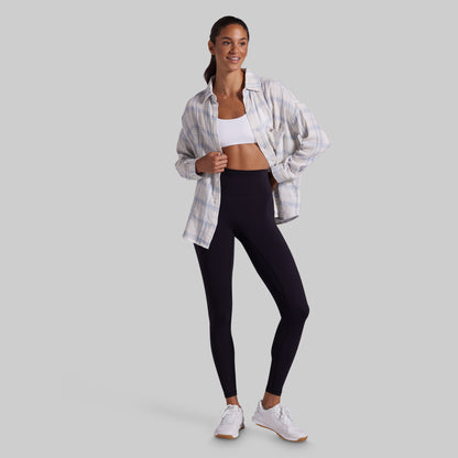 A woman modeling a pair of black high-waisted leggings, styled with an open white and blue plaid button-down shirt, a white sports bra, and white sneakers, set against a solid gray studio background.