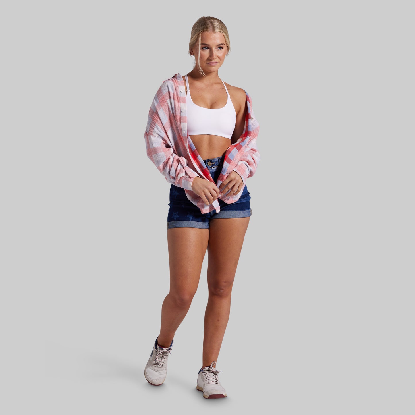 A woman modeling a pink and white plaid button-up shirt worn open over a white sports bra and dark blue denim shorts, standing against a solid gray studio background.
