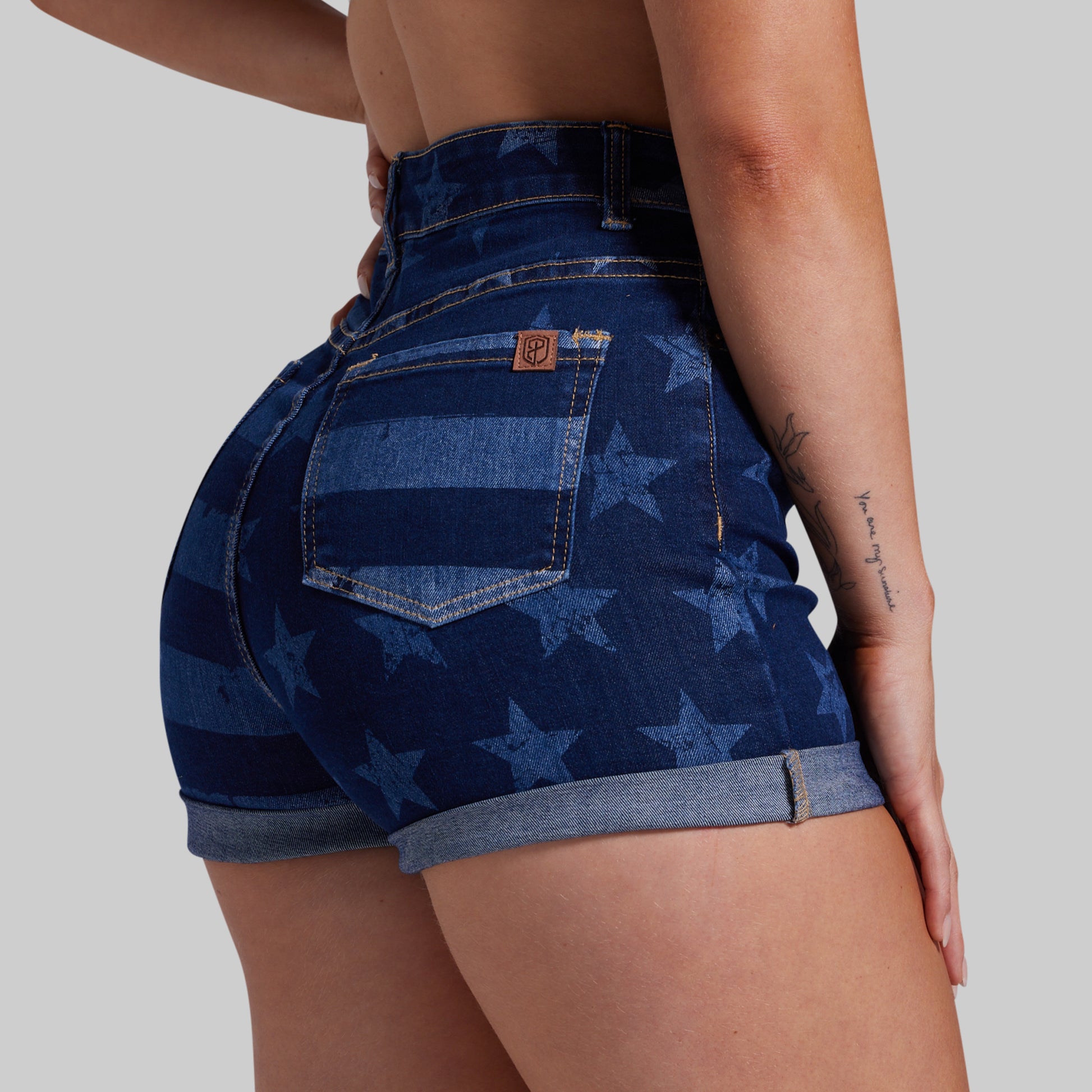 Close-up back view of a model wearing high-waisted denim shorts with a blue and navy American flag pattern featuring stars and stripes, on a solid gray background.