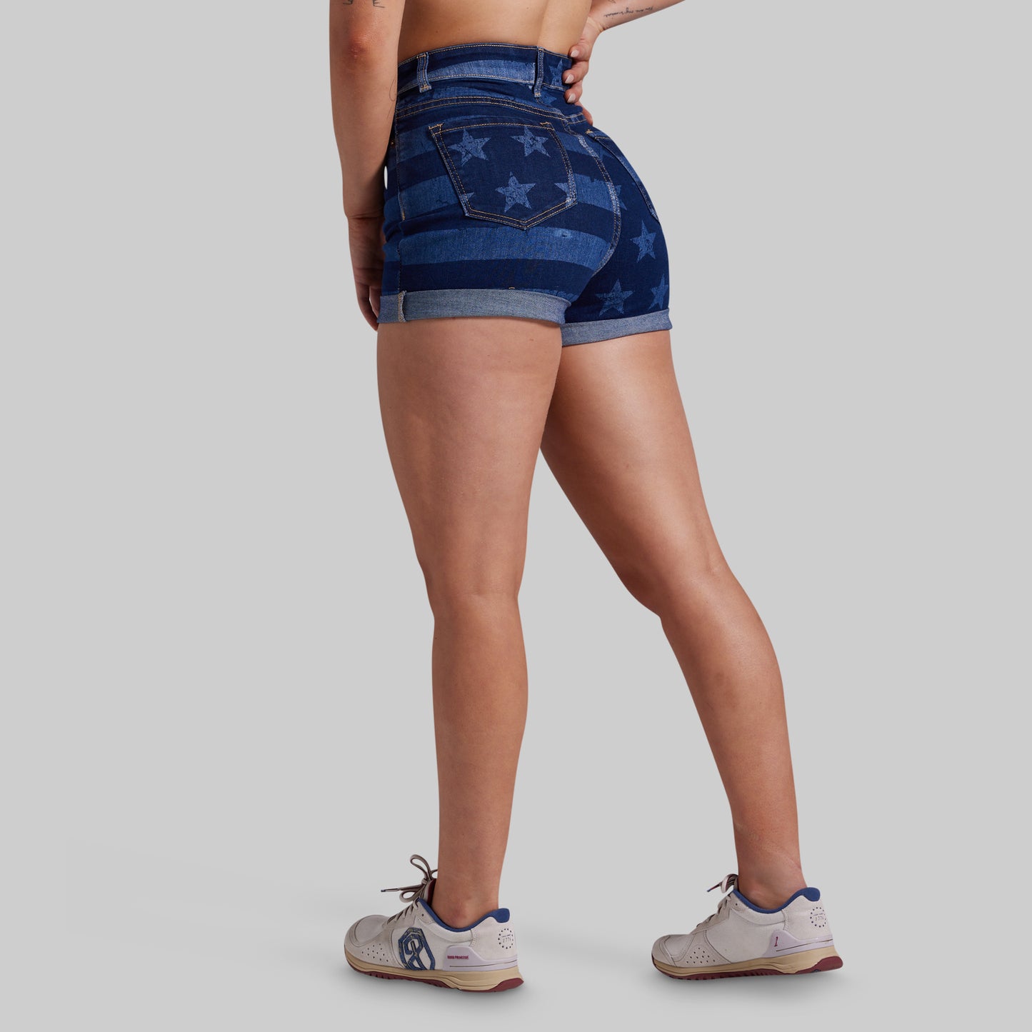 Rear three-quarter view of a model wearing high-waisted blue denim shorts with a faded star pattern, paired with white athletic sneakers, against a solid light gray background.