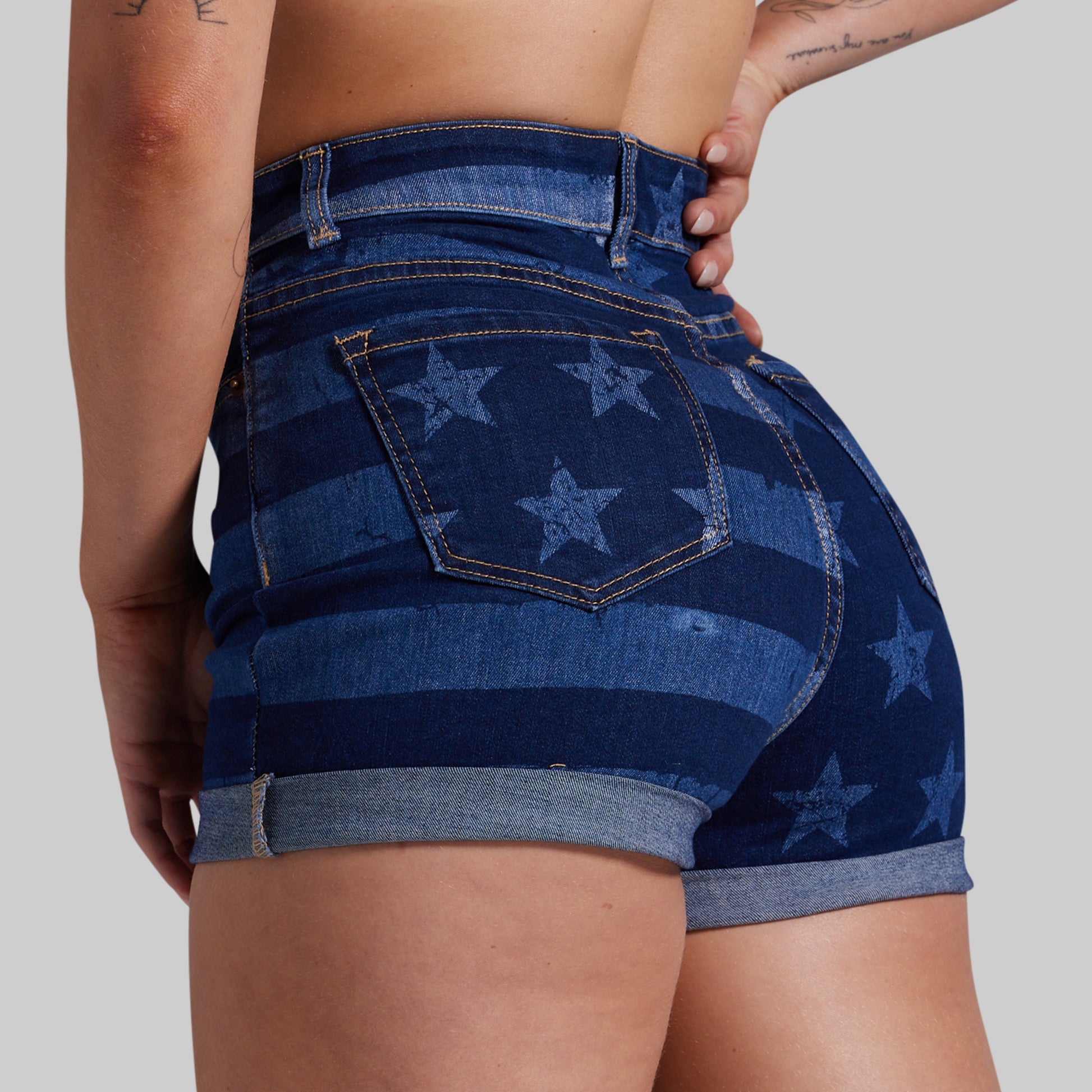 Close-up back view of a person wearing high-waisted denim shorts with a blue and navy star-and-stripe pattern, shown against a light gray studio background.