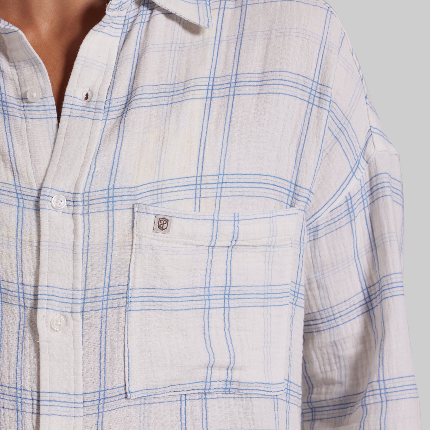 Close-up of a person wearing a long-sleeve button-up shirt in off-white with a blue plaid pattern and a chest pocket featuring a small shield logo.