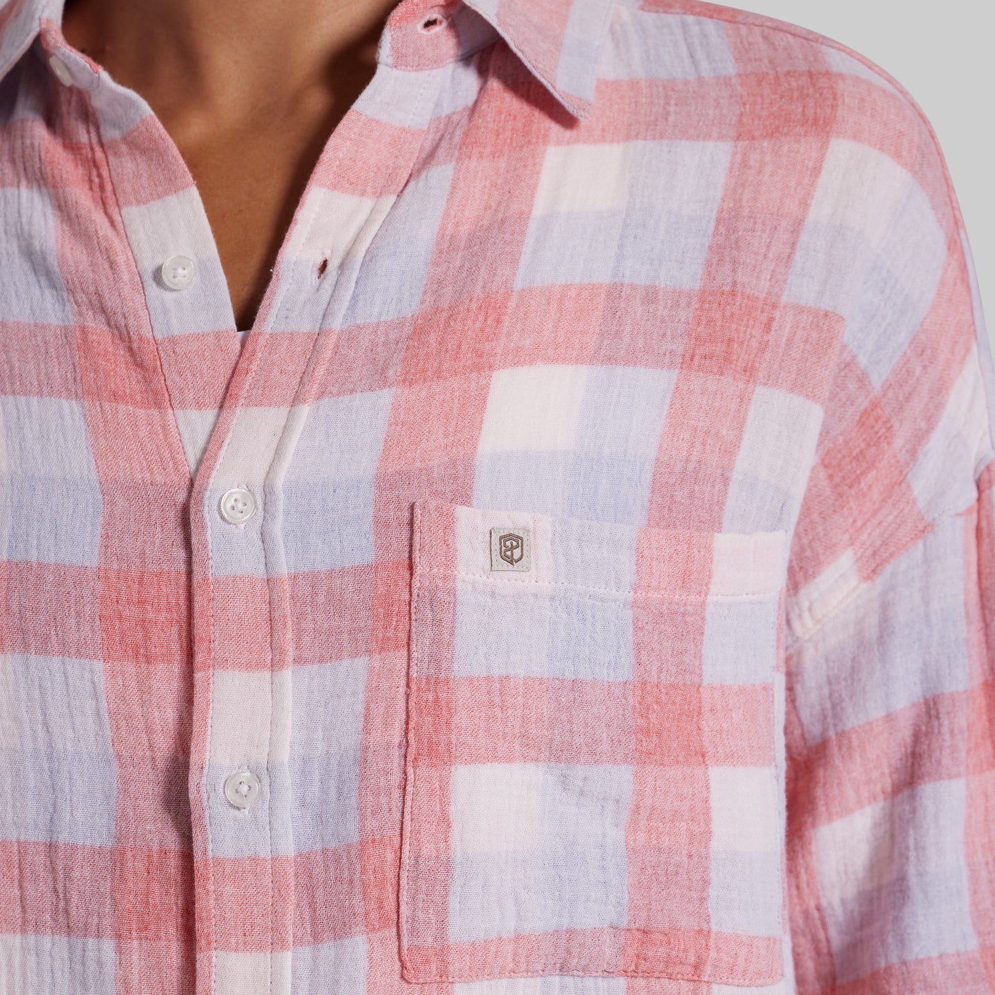 A close-up of a person wearing a long-sleeve button-up shirt with a pink, white, and light blue plaid pattern. The shirt features a single chest pocket with a small shield logo patch on the left chest.