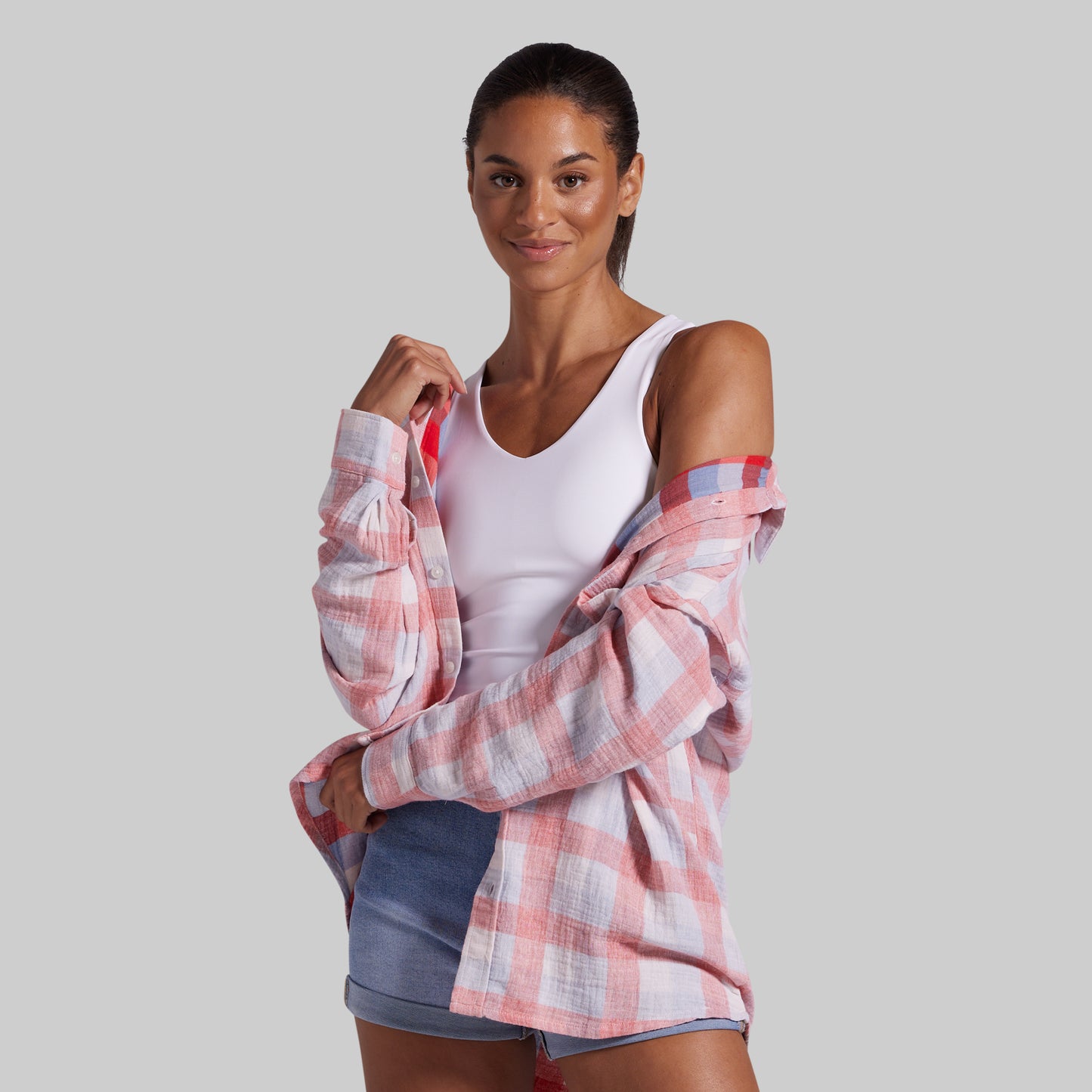 A woman modeling a pink and white plaid flannel shirt with cold-shoulder styling over a white tank top and denim shorts, set against a solid gray background.