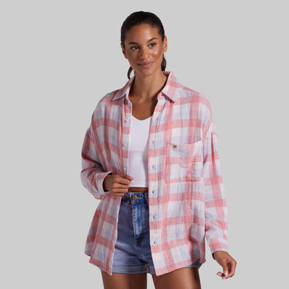 A woman modeling a pink and white plaid button-up shirt over a white tank top and denim shorts, set against a solid gray studio background.