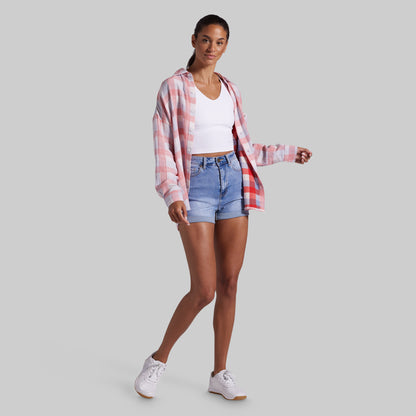 A woman modeling a pink and white plaid button-up shirt over a white crop top and light blue denim shorts, paired with white sneakers, against a solid gray background.