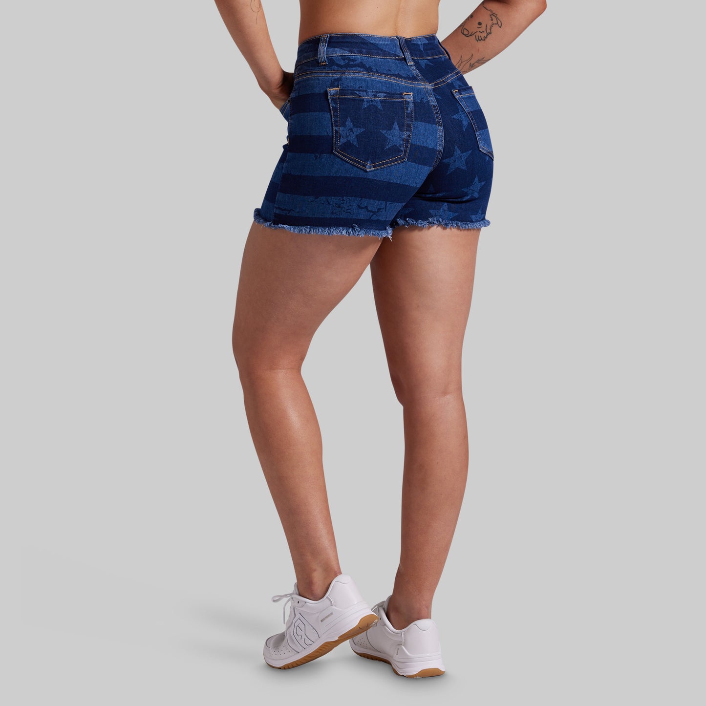 A model from the back wearing mid-rise, frayed-hem denim shorts with horizontal dark and light blue stripes and a subtle star pattern, paired with white sneakers on a solid gray background.