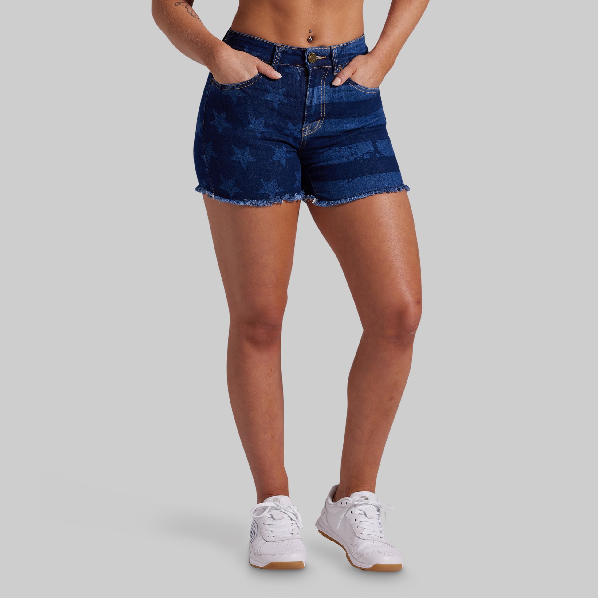 A model wearing dark blue denim shorts with a tonal star pattern and frayed hems, paired with white sneakers, shown from the waist down against a solid gray background.