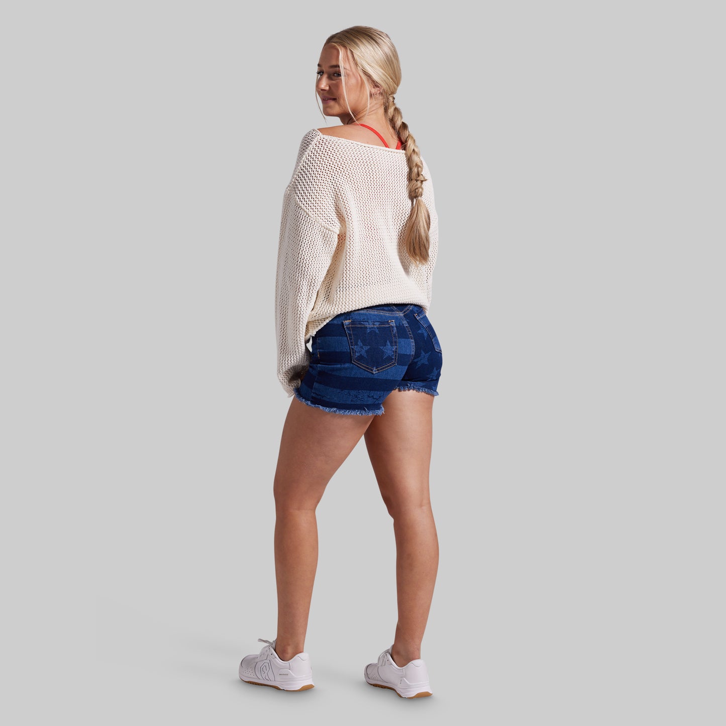 A woman is shown from the back wearing a cream knit sweater, blue denim shorts, and white sneakers against a solid gray background.